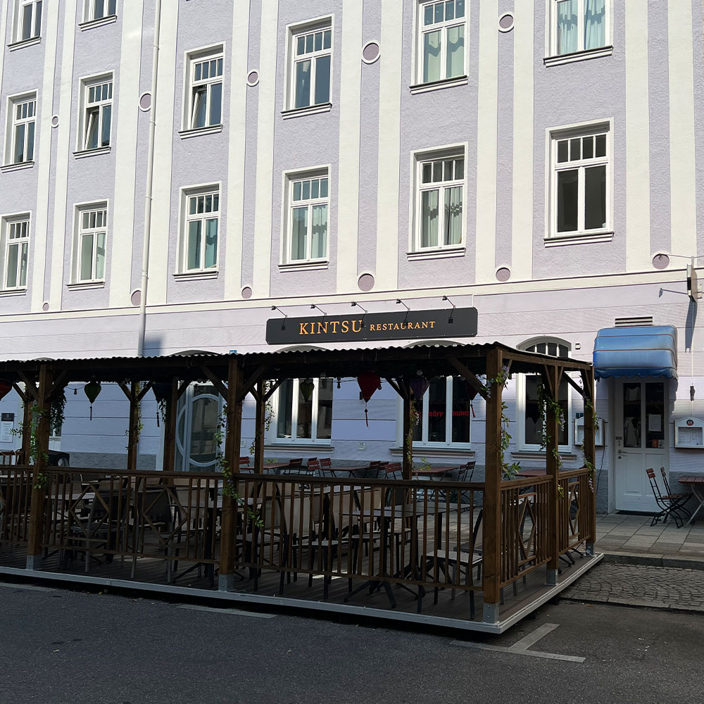 Ein leeres Straßencafé mit Holzmöbeln vor einem pastellfarbenen Gebäude.