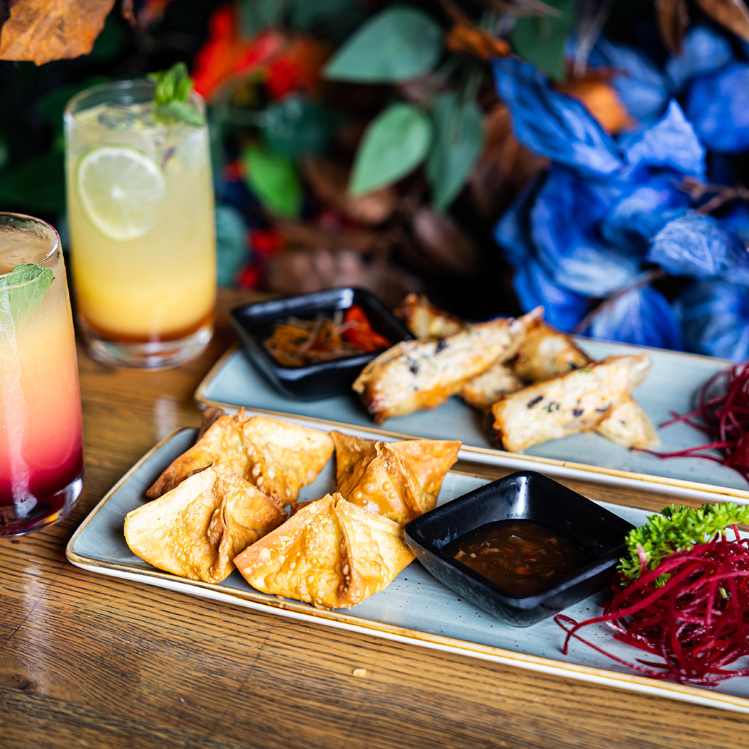Bunte asiatische Snacks und Dips mit zwei farbigen Cocktails auf Holztisch