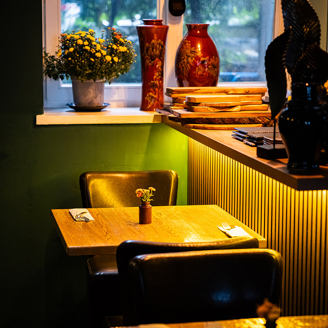 Ein leerer Holztisch mit zwei braunen Stühlen steht nahe einem Fenster mit Blumen und roten Vasen