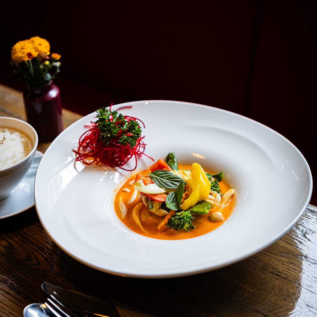 Bunter Gemüseeintopf in roter Sauce auf weißem Teller mit Reis und Blumenarrangement