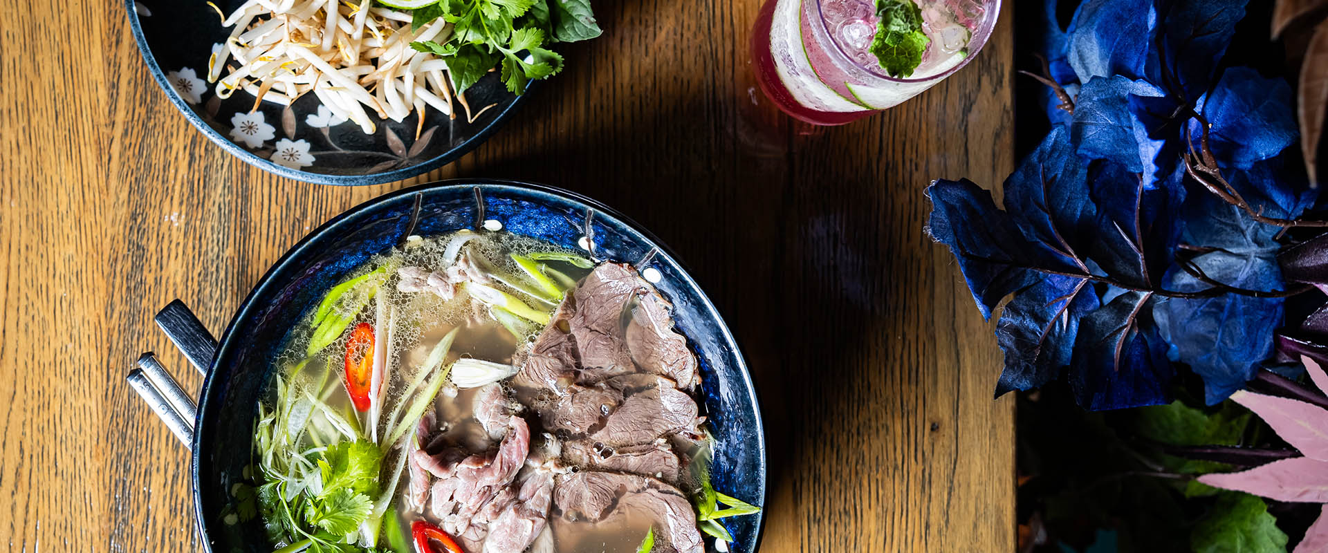 Eine Schale mit Pho Suppe und Rindfleischscheiben auf Holz, daneben frische Kräuter