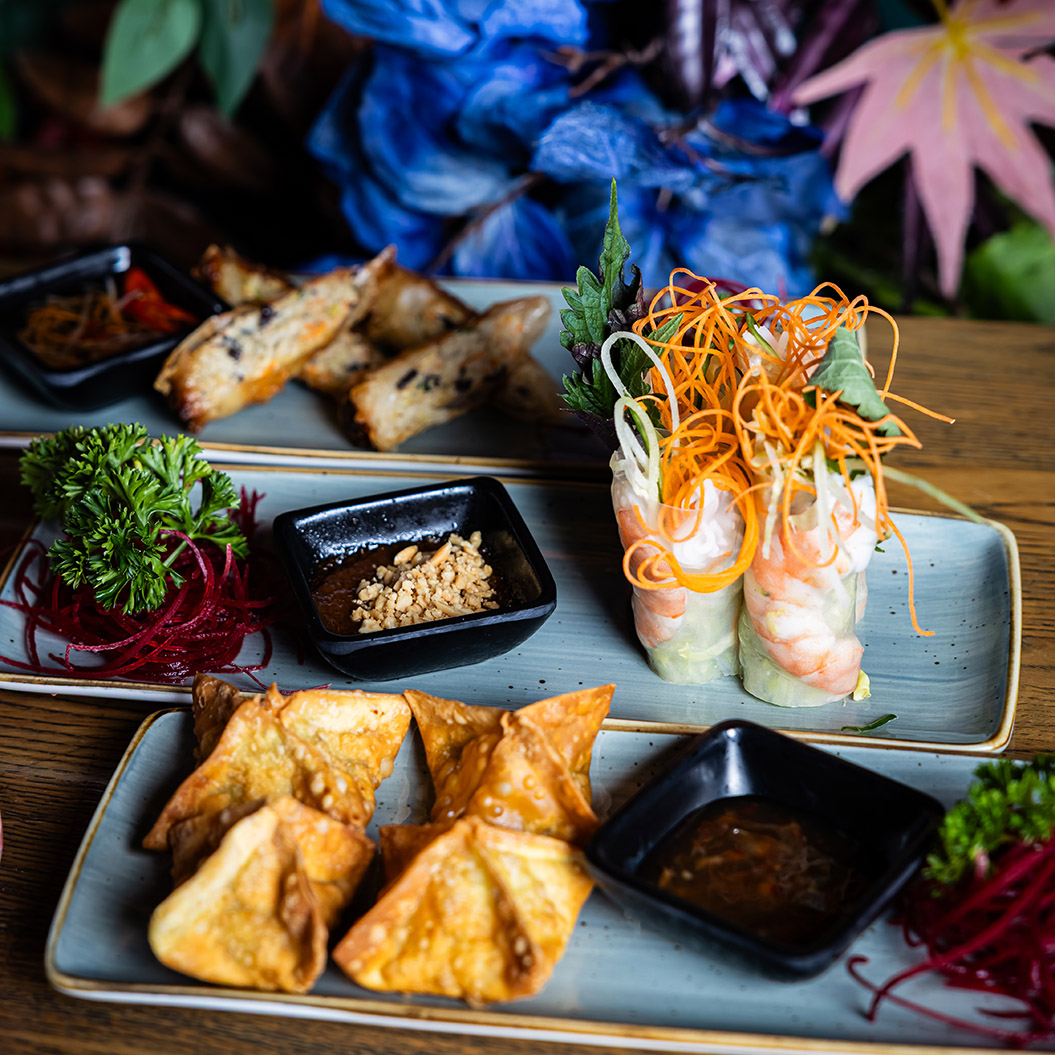 Verschiedene asiatische Vorspeisen mit Gemüse, gebratenen Teigtaschen und Frühlingsrollen auf stilvoll angerichteten Platten, umgeben von dekorativen Pflanzen und bunten Blüten
