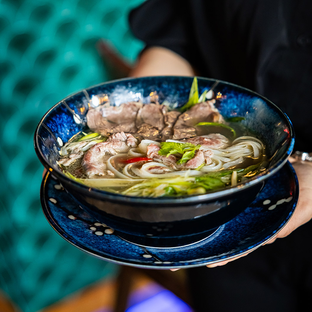Eine Person hält eine große blaue Schale mit dampfender Pho Suppe und frischen Kräutern